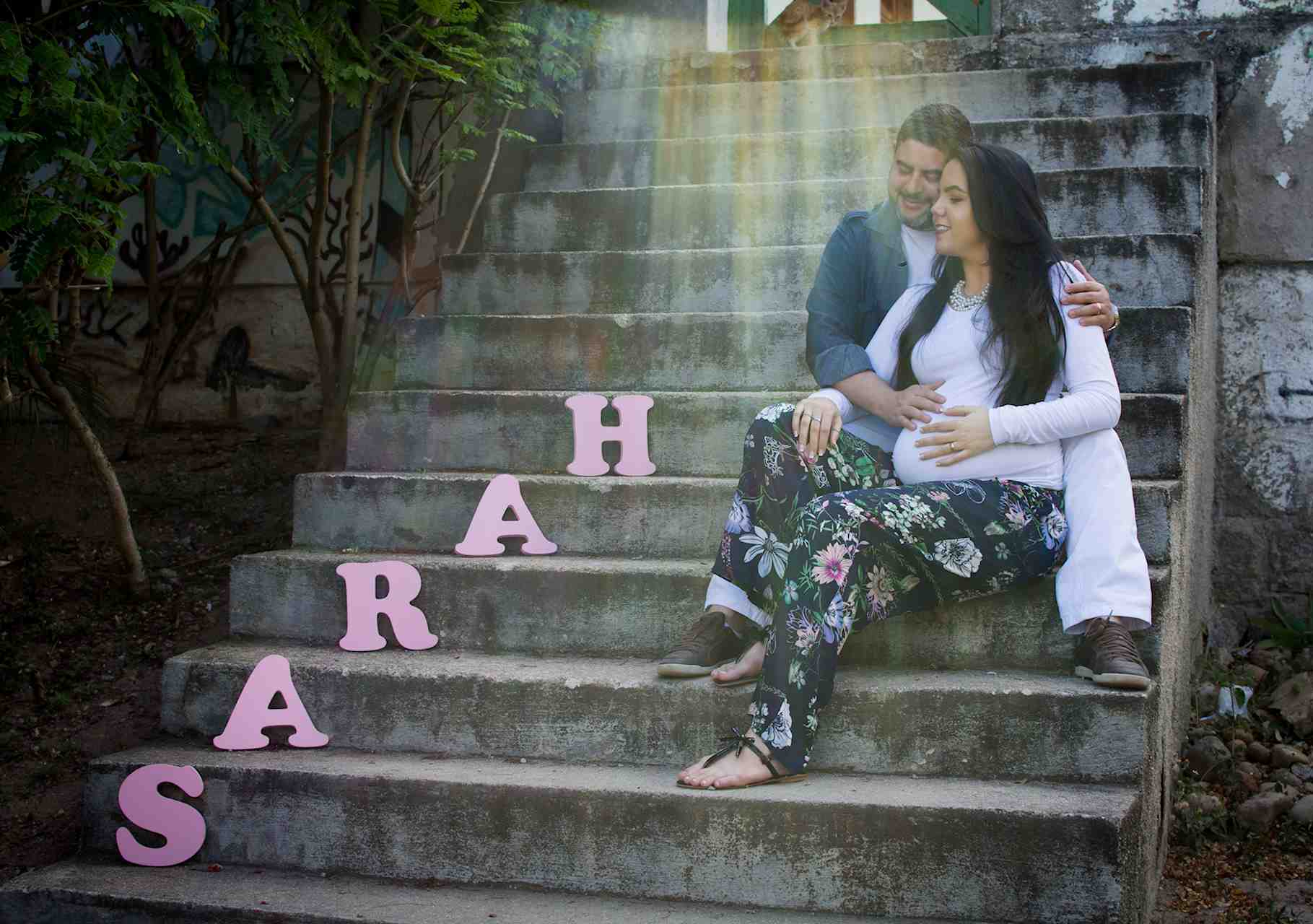Retrato de Hélio e Ceiça, sentados em degraus, ela grávida, e ambos olhando as letras do nome da filha que esperam, Sarah